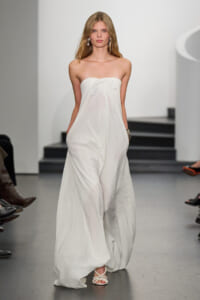 Model walking a runway in a strapless white maxi dress with a front bow and pockets, wearing dangling earrings and braided sandals.