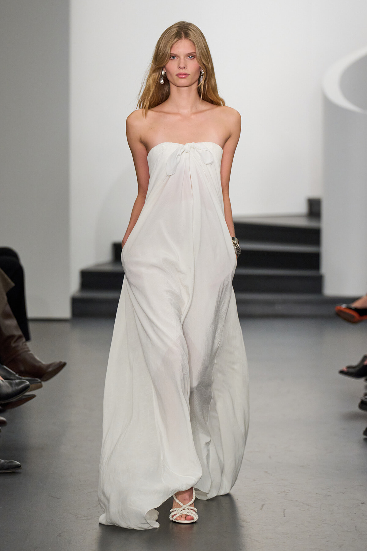 Model walking a runway in a strapless white maxi dress with a front bow and pockets, wearing dangling earrings and braided sandals.