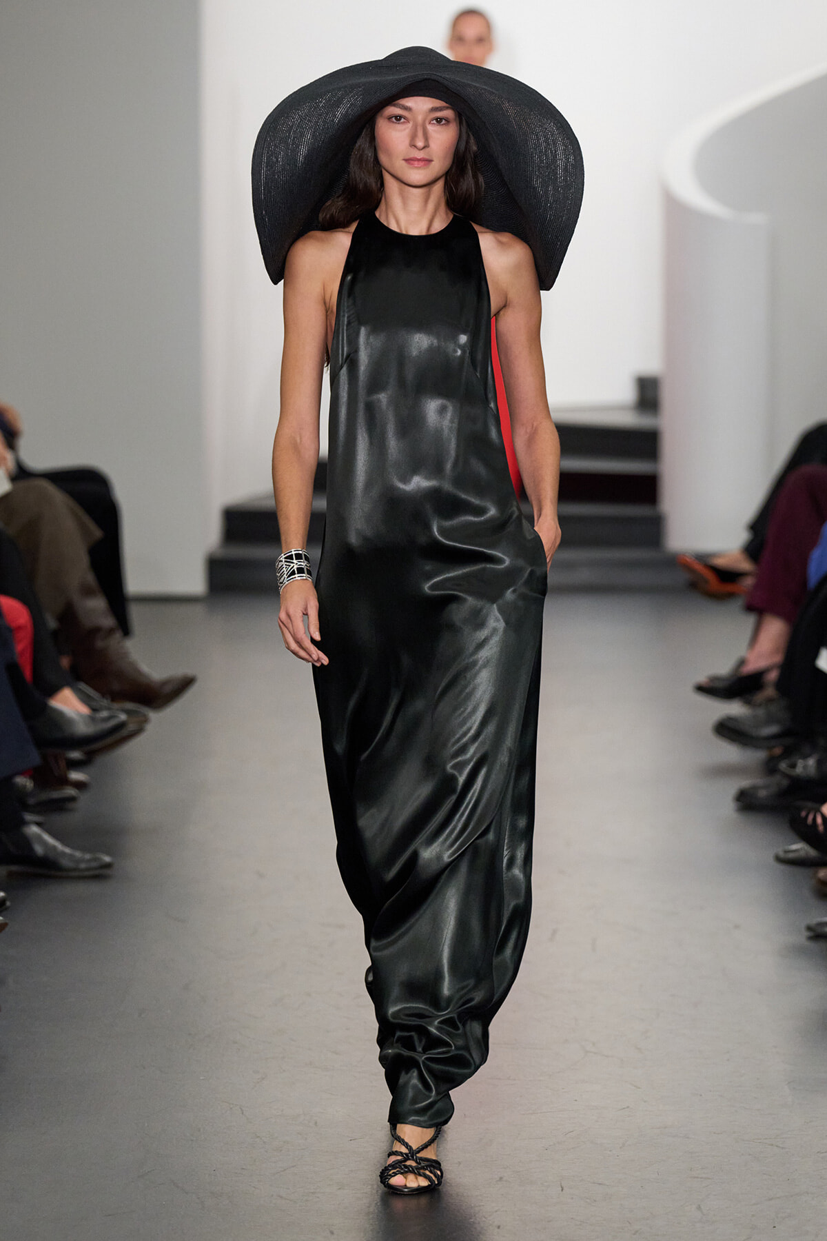 Model walking a fashion runway in a long black satin halter dress with a oversized black brimmed hat.