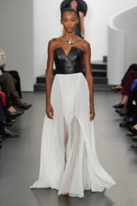 Model walking a fashion runway in a black strapless leather bodice and a long white pleated chiffon skirt, with a statement silver necklace.