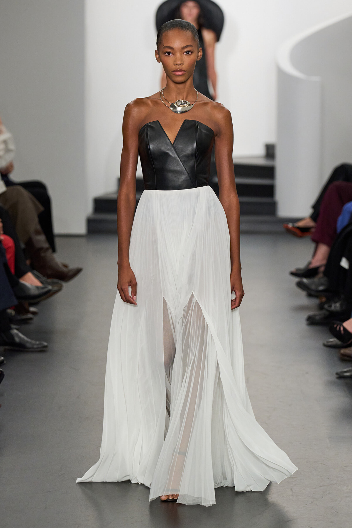 Model walking a fashion runway in a black strapless leather bodice and a long white pleated chiffon skirt, with a statement silver necklace.