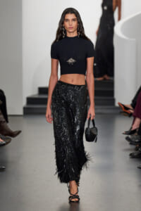 Model walks a runway in a black cropped top and high-waisted sequined pants, holding a small black handbag and wearing statement jewelry.