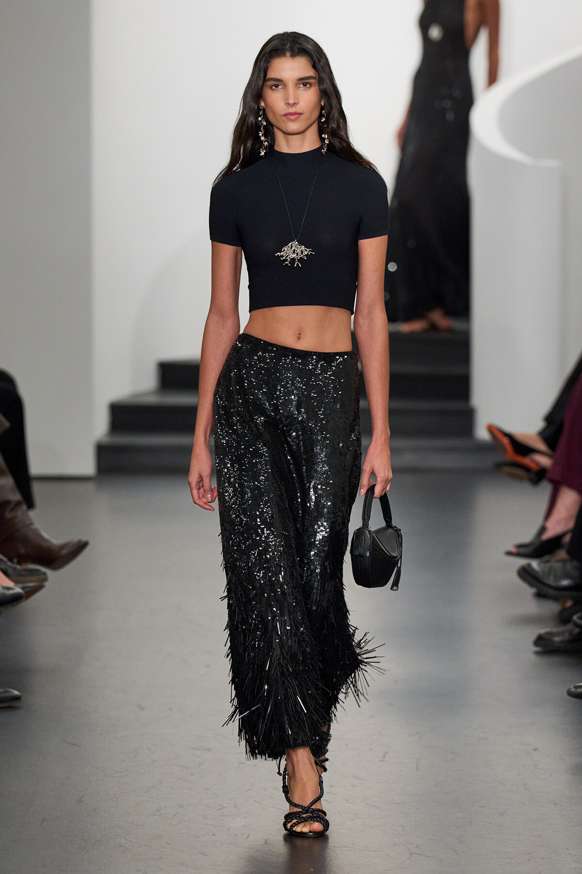 Model walks a runway in a black cropped top and high-waisted sequined pants, holding a small black handbag and wearing statement jewelry.