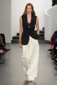 Model walking a runway in a black sleeveless blazer and wide white pants, accessorized with a bold necklace and bracelet.