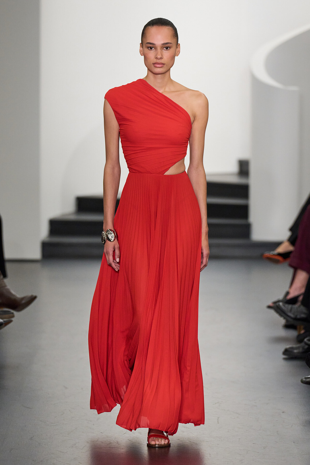 Model walking a runway in a bright red one-shoulder crop top and matching pleated maxi skirt.
