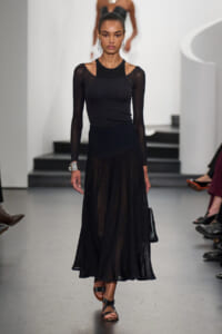 Model walking the runway in a black ensemble: mesh-sleeve top and sheer, pleated midi skirt, with statement earrings and sandals.