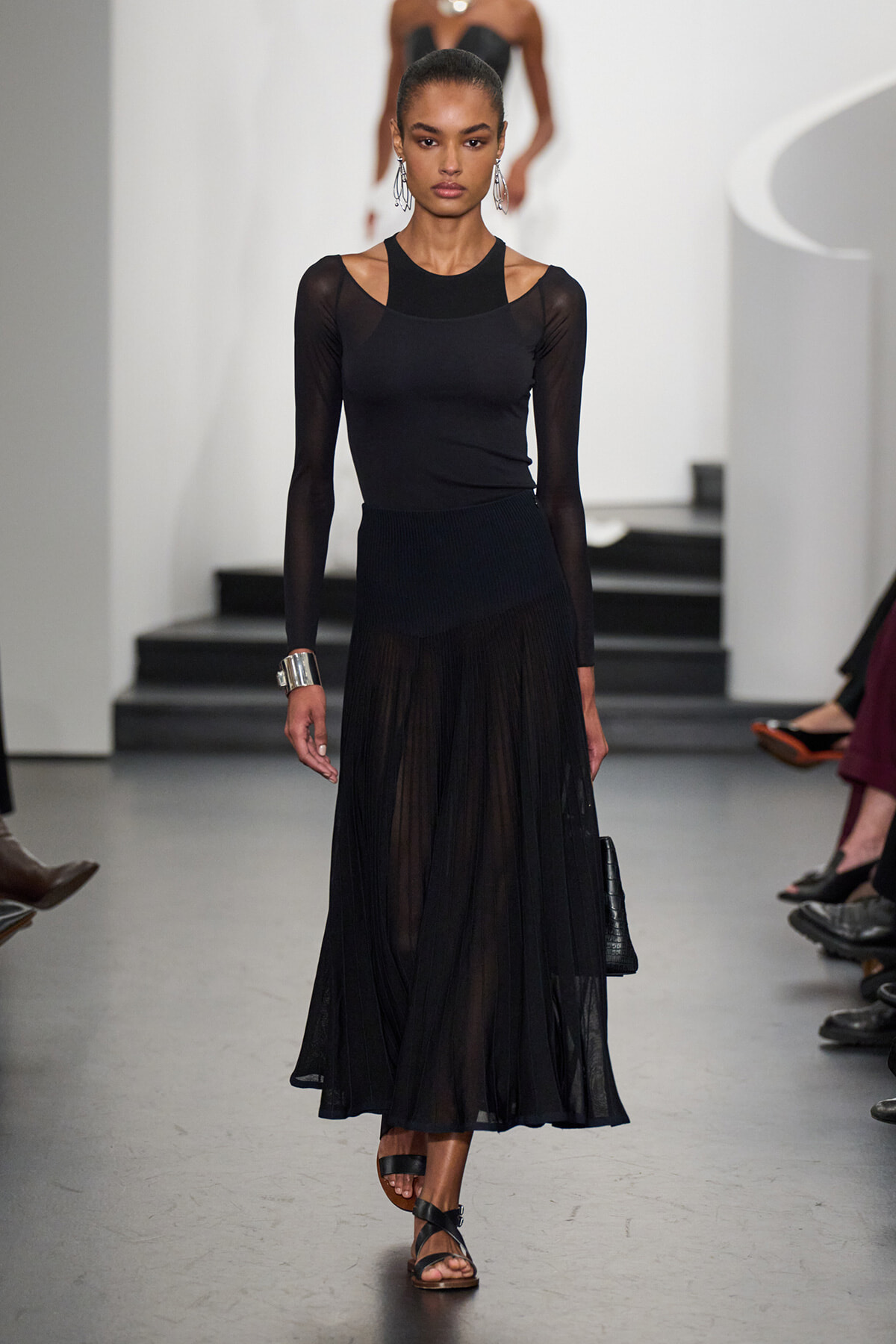Model walking the runway in a black ensemble: mesh-sleeve top and sheer, pleated midi skirt, with statement earrings and sandals.