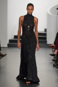 Model on a runway wearing a sleeveless black turtleneck gown with long fringe strands and a pendant necklace.