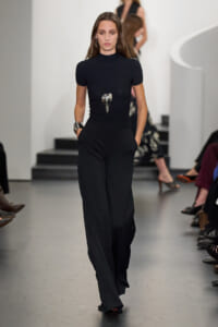 Model walking runway in a fitted black turtleneck and wide-leg black pants, holding hands in pockets, with a bold pendant necklace and chunky silver bracelets.