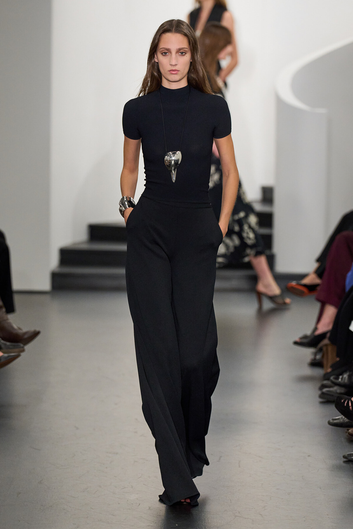 Model walking runway in a fitted black turtleneck and wide-leg black pants, holding hands in pockets, with a bold pendant necklace and chunky silver bracelets.
