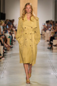 Model walking the runway in a pale yellow perforated trench coat with a waist belt and double-breasted buttons.