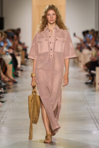 Model on a runway wearing a pastel pink perforated shirt-dress with large front pockets and dark buttons, carrying a tan fringe handbag and wearing tan sandals.