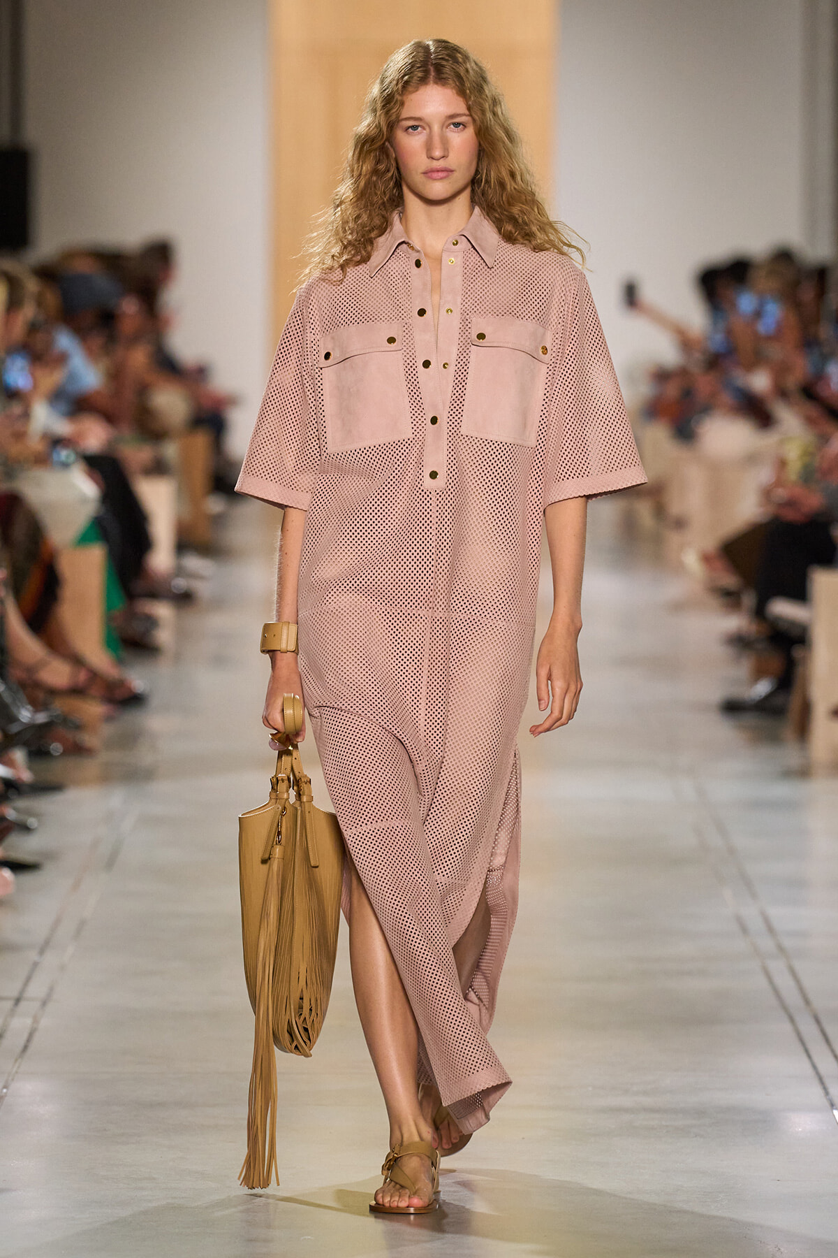 Model on a runway wearing a pastel pink perforated shirt-dress with large front pockets and dark buttons, carrying a tan fringe handbag and wearing tan sandals.