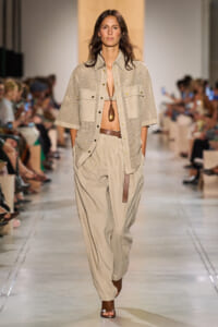 Model walks the runway in a beige perforated short-sleeve shirt, matching wide trousers, brown belt, and a long pendant necklace.