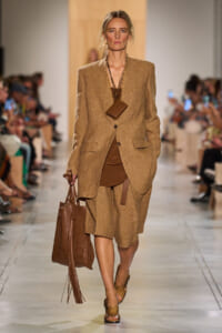 Model walking runway in a tan/khaki oversized blazer and matching shorts with brown top, carrying a large brown tote bag and a small brown pouch necklace.