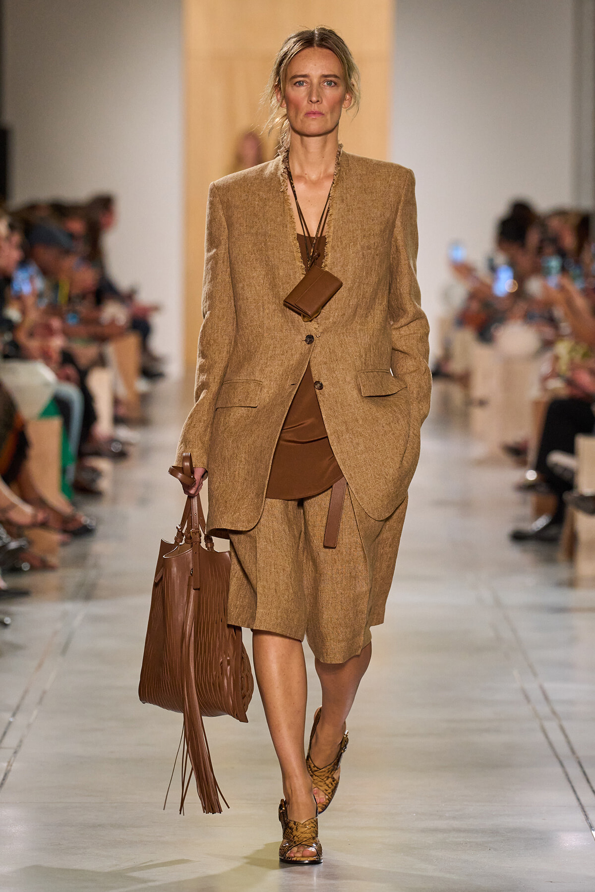 Model walking runway in a tan/khaki oversized blazer and matching shorts with brown top, carrying a large brown tote bag and a small brown pouch necklace.