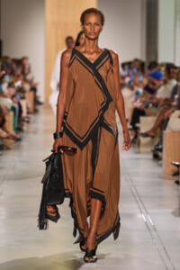 Model walking a brown, flowing dress with bold black geometric trim on a runway, carrying a black fringed handbag.