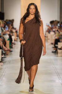 Model walks the runway in a brown draped one-shoulder dress, carrying a fringed brown handbag.