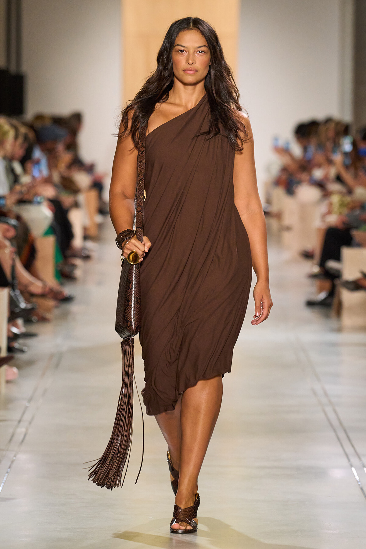 Model walks the runway in a brown draped one-shoulder dress, carrying a fringed brown handbag.