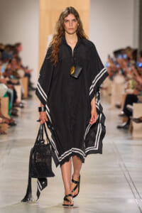 Model walking a runway in a black poncho with white striped trim, carrying a black woven bag and wearing flat sandals.