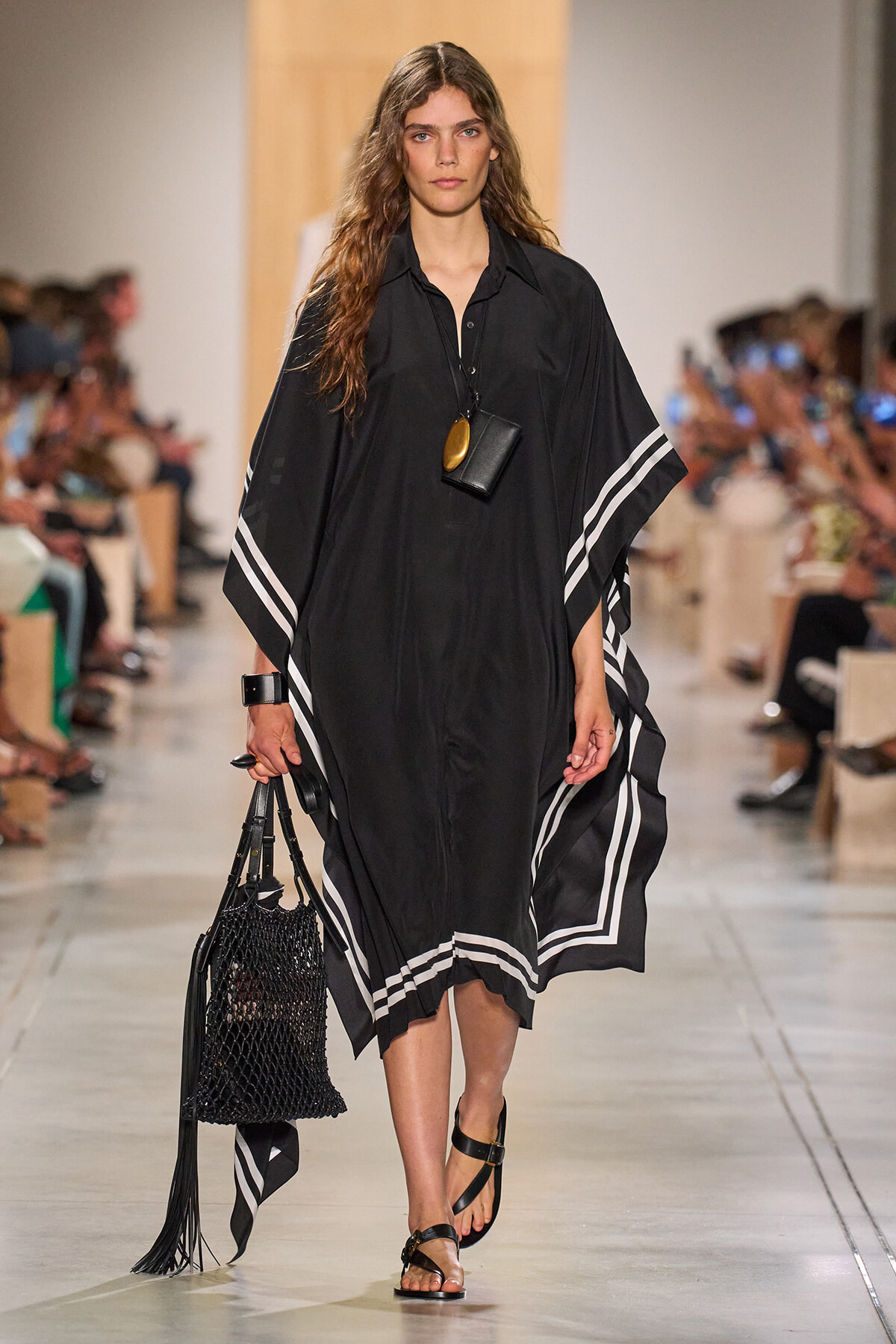 Model walking a runway in a black poncho with white striped trim, carrying a black woven bag and wearing flat sandals.