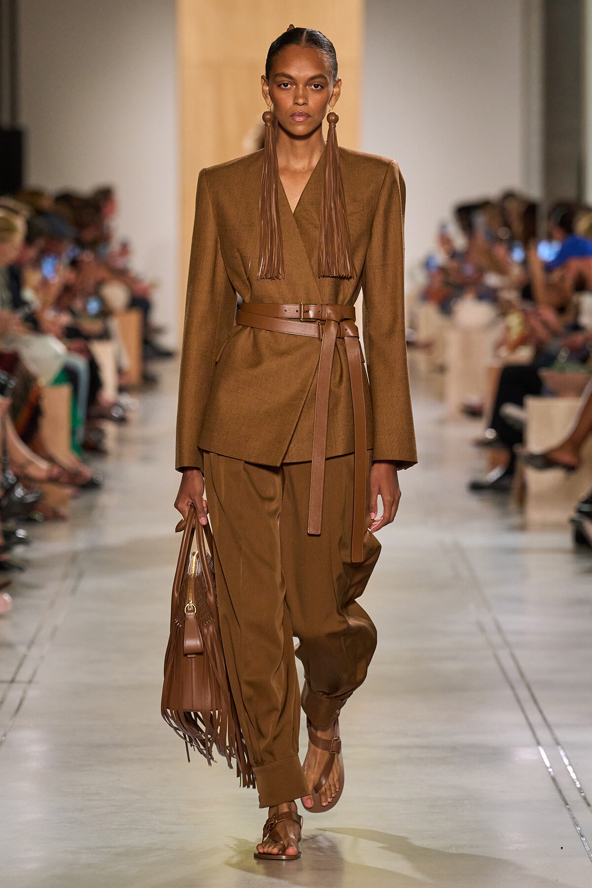 Model walks down runway in a brown tailored suit with a belt, matching pants, and fringe earrings; carries a brown fringed handbag.