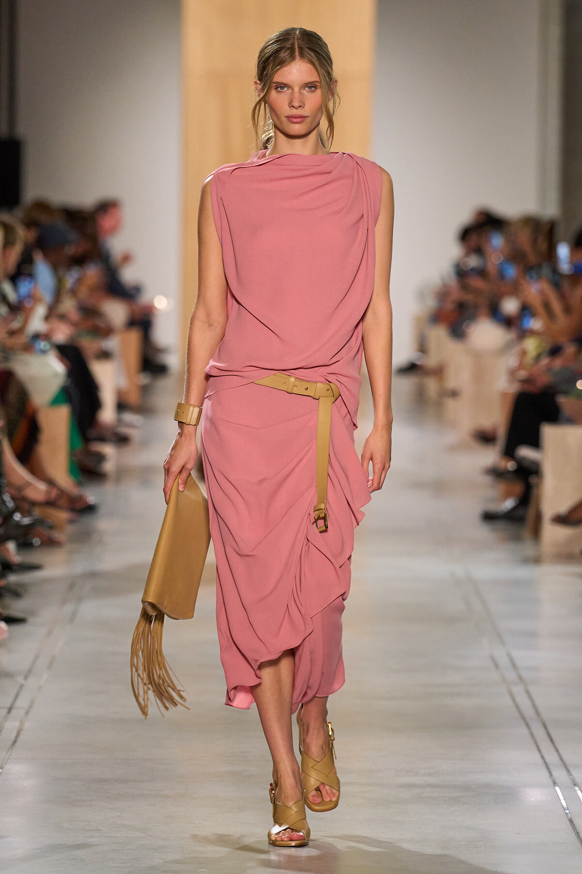 Model walking a pink draped dress on a runway with a tan belt and tan fringe handbag; audience seated on either side.