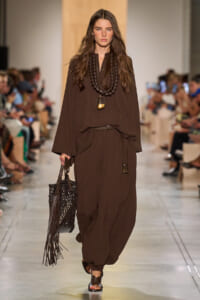 Model walks down runway in all-brown ensemble: loose cape-like top, pleated harem pants, chunky beaded necklace, and a fringed brown bag.