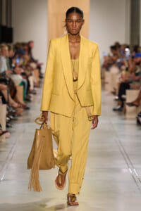Model walking a runway in a lemon-yellow blazer and matching wide-leg pants, carrying a tan fringed tote.