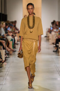Model walks the runway in a camel-toned outfit: oversized top, draped midi skirt, and a chunky beaded necklace with a large pendant.