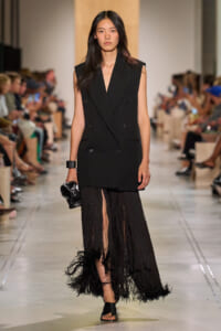 Model walks the runway in a sharp black sleeveless double-breasted blazer paired with a long black fringe skirt with feathered hem, clutch in hand.