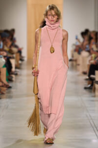 Model walking a runway in a pale pink sleeveless knit turtleneck dress with matching wide pants, carrying a tan fringe bag and wearing a long pendant necklace.