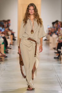 Model walking the runway in a loose beige jumpsuit with a brown belt and matching sandals, accessorized with a two-tone pendant necklace on the catwalk locale.