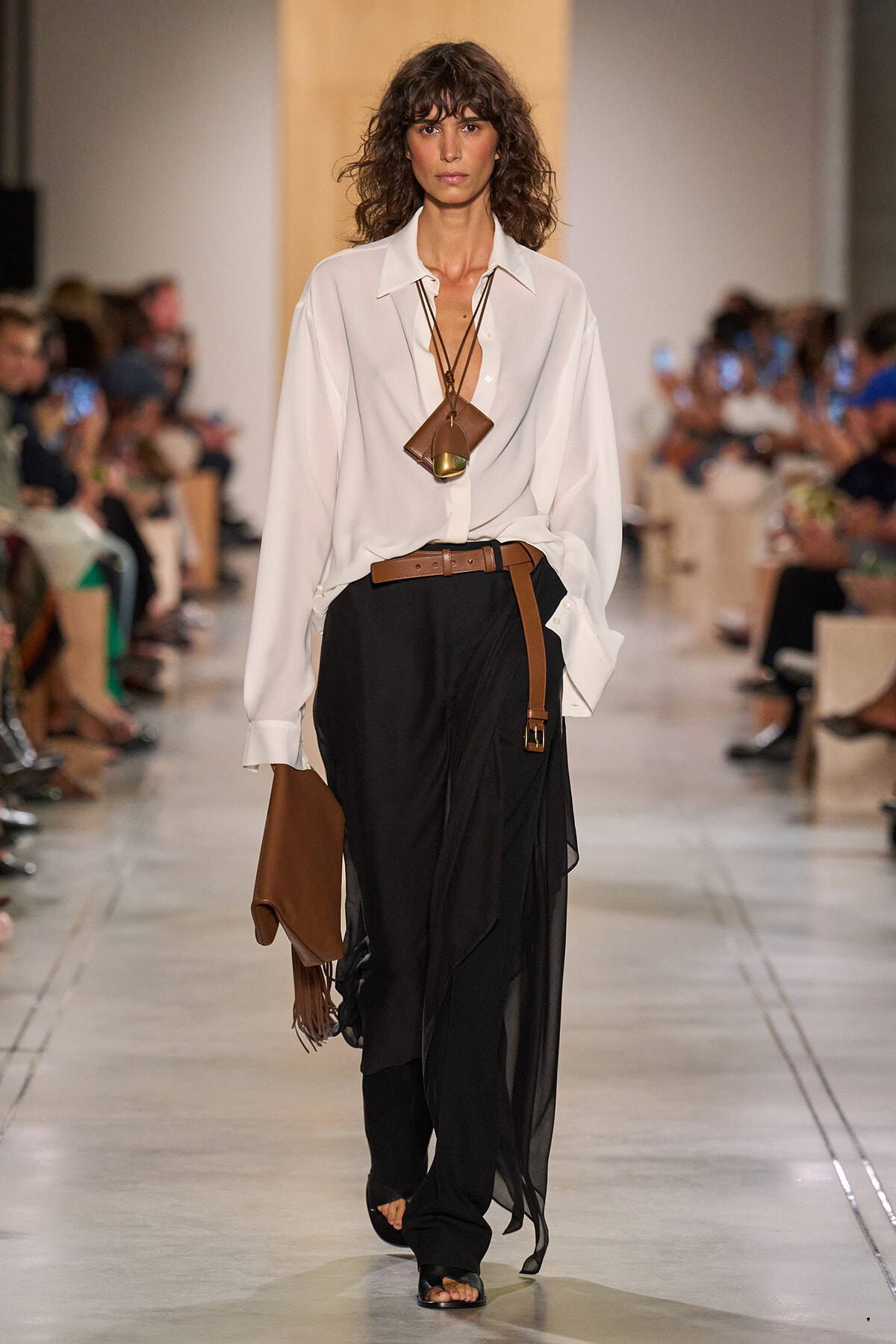 Model on runway wearing a white blouse tucked into a long black draped skirt with a brown belt, tan bag, and layered brown necklaces.