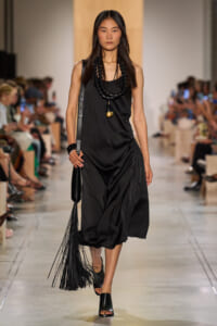 Models black sleeveless midi dress with draped side, accessorized by stacked black beads and a long pendant, walking a runway with a fringed black bag