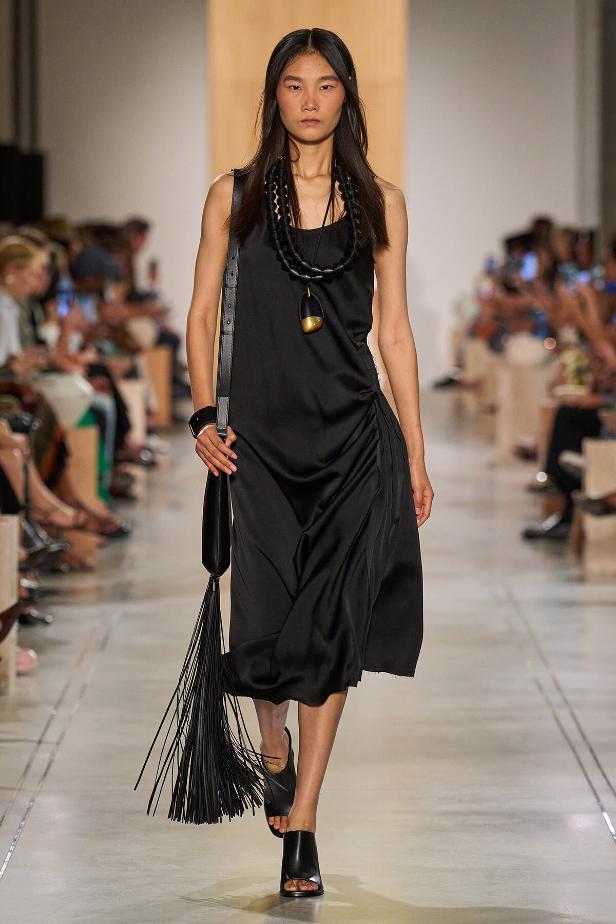 Models black sleeveless midi dress with draped side, accessorized by stacked black beads and a long pendant, walking a runway with a fringed black bag