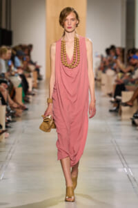Model walks the runway in a dusty pink, draped midi dress with a chunky wooden bead necklace and tan sandals, holding a small tan clutch.