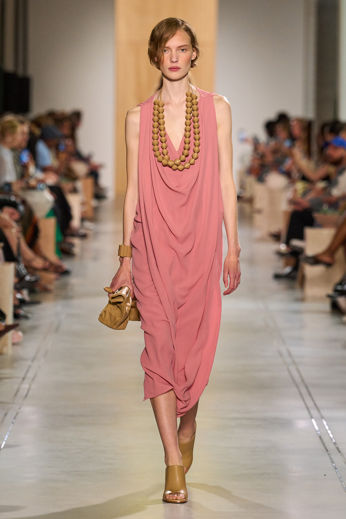 Model walks the runway in a dusty pink, draped midi dress with a chunky wooden bead necklace and tan sandals, holding a small tan clutch.