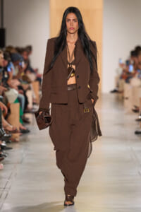 Model on a runway wearing a brown suit with a structured blazer and draped, loose pants, black bralette visible, layered necklace, and a brown textured clutch.