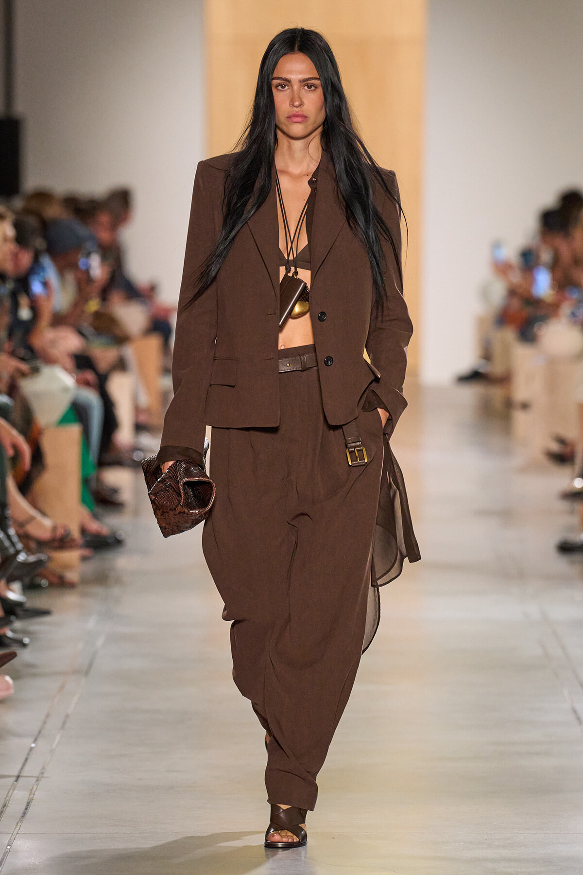 Model on a runway wearing a brown suit with a structured blazer and draped, loose pants, black bralette visible, layered necklace, and a brown textured clutch.