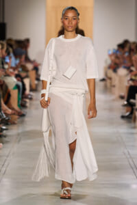 Model on a fashion runway wearing an all-white outfit: sheer knit top, flowing asymmetric skirt with a belt, and white accessories including a small pendant bag and a fringed tote with sandals.