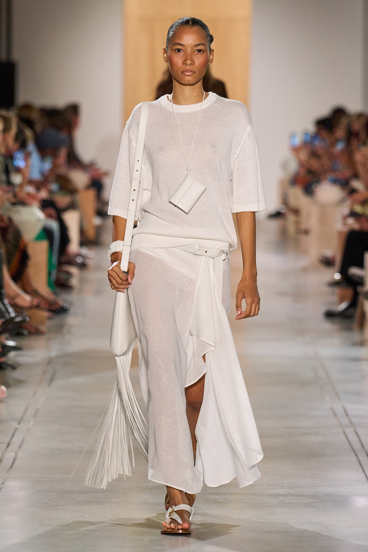 Model on a fashion runway wearing an all-white outfit: sheer knit top, flowing asymmetric skirt with a belt, and white accessories including a small pendant bag and a fringed tote with sandals.