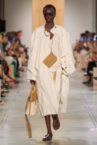 Model walks a fashion runway in an oversized cream trench with a beige mini bag hanging from a long strap and brown sandals, wearing dark sunglasses.