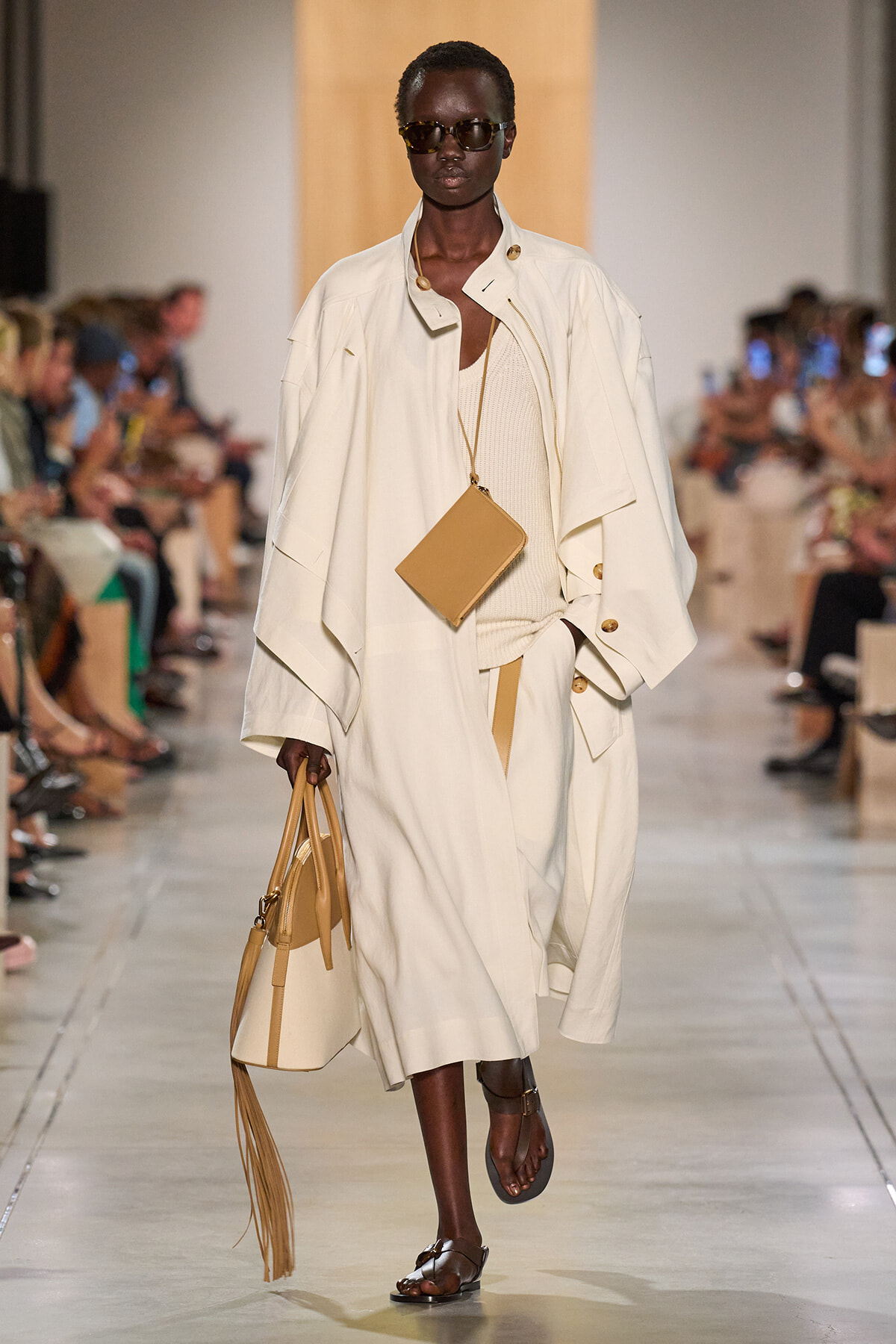 Model walks a fashion runway in an oversized cream trench with a beige mini bag hanging from a long strap and brown sandals, wearing dark sunglasses.