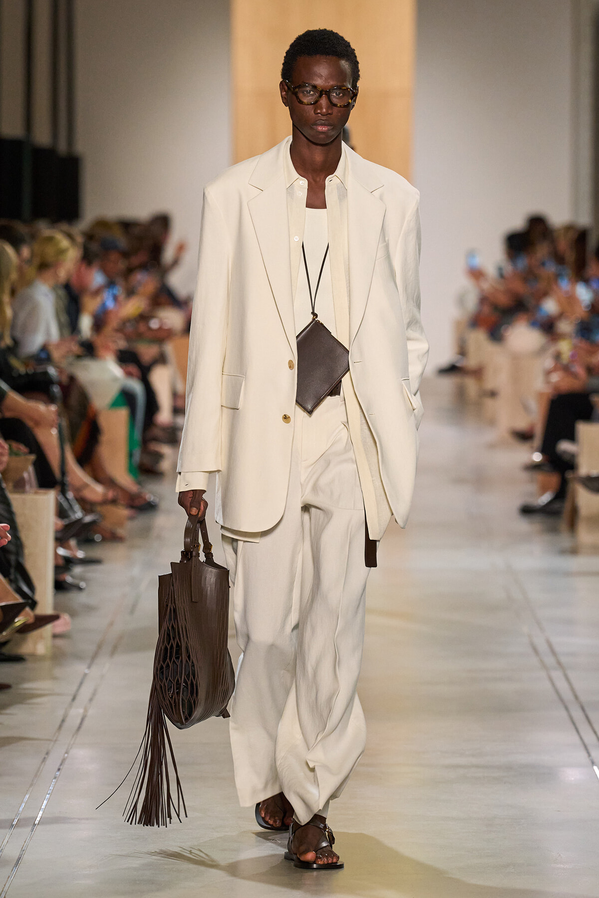 Model walks the runway in an oversized cream suit with a white shirt, carrying a brown fringed tote and a small brown pouch necklace.