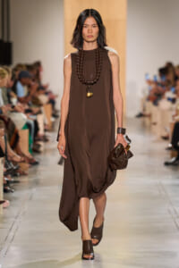 Model walks a runway in a loose brown sleeveless dress with a chunky beaded necklace and brown sandals, carrying a brown clutch.