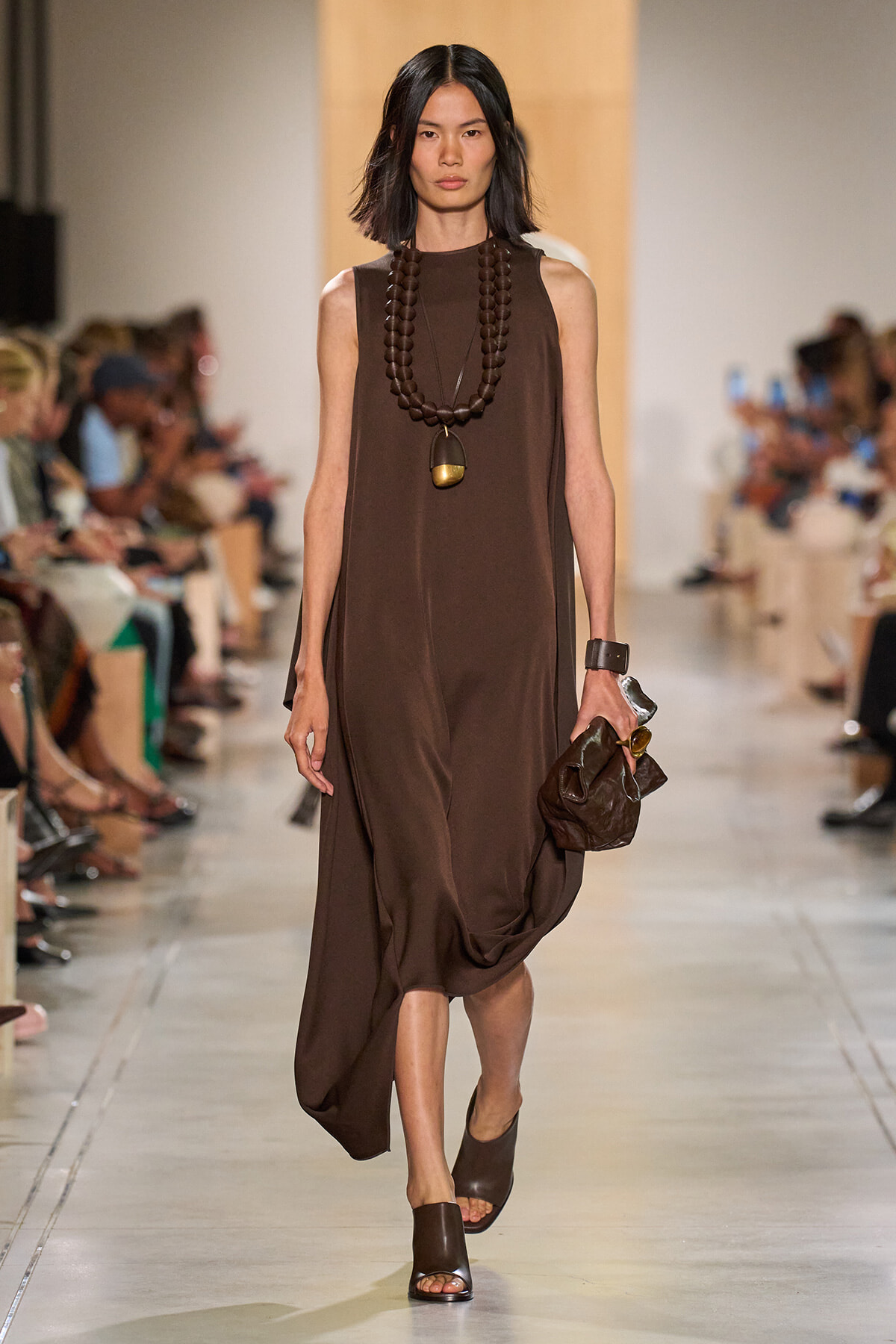 Model walks a runway in a loose brown sleeveless dress with a chunky beaded necklace and brown sandals, carrying a brown clutch.