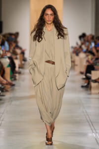 Model walking a runway in a loose beige suit with draped top, brown belt, and brown sandals.