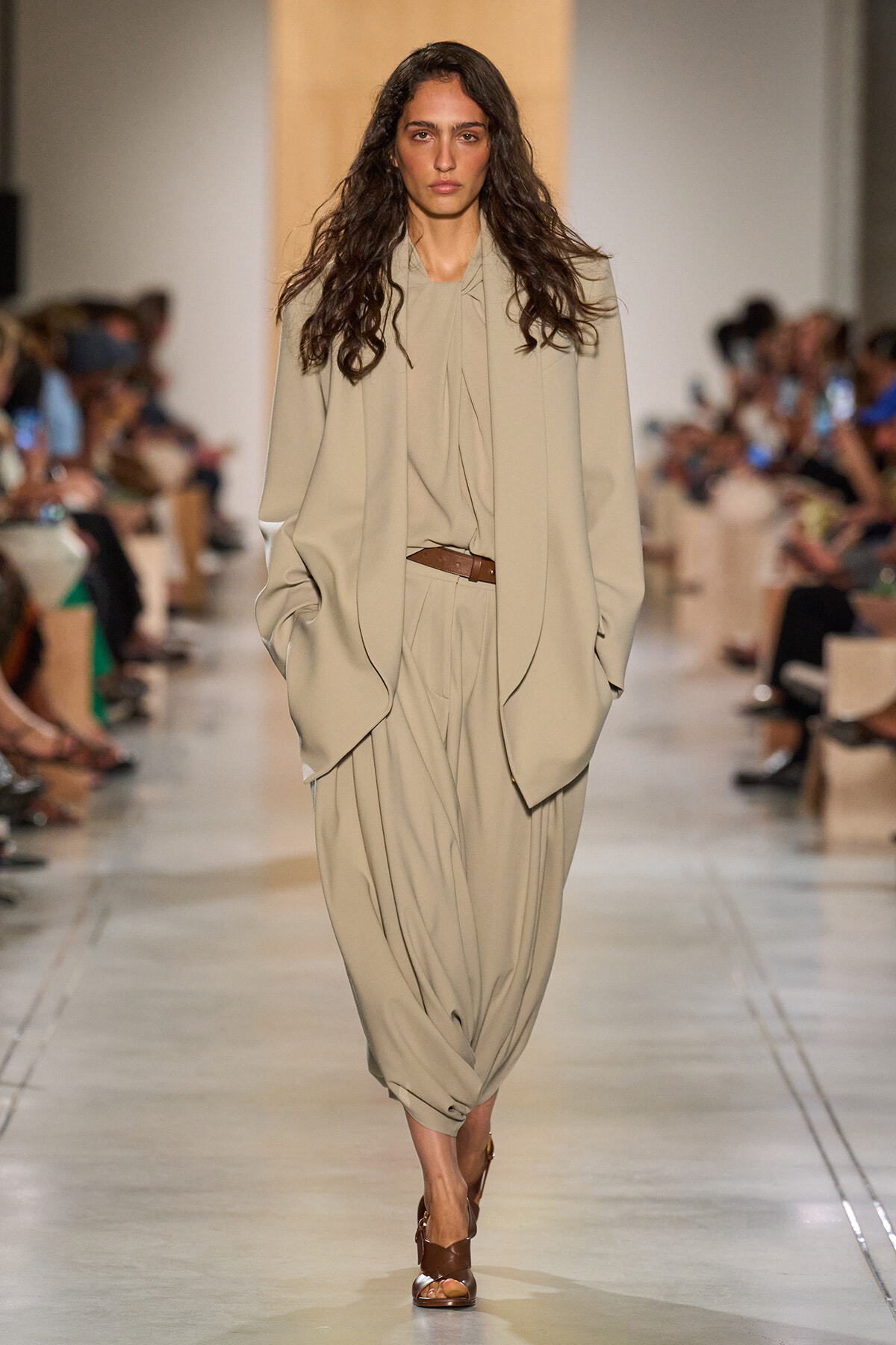 Model walking a runway in a loose beige suit with draped top, brown belt, and brown sandals.