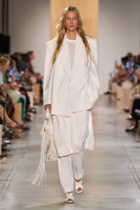 Model walks the runway in an oversized white double-breasted blazer and wide-leg white pants, carrying a white netted bag with fringe.